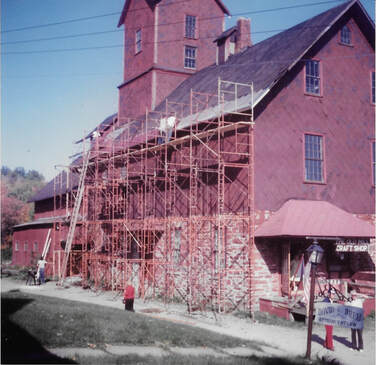 Picture 12 – New shingles being installed on the parking lot side of the main mill building by David Billado and crew, October 6, 1982.  Unfortunately, plastic sheeting was used as an underlayment for the shingles, and this quickly deteriorated, ultimately forcing the removal of these shingles so that proper underlayment could be installed.  Note the sign in the lower right of the picture for David Drew when he had his law office in the first floor of the mill house.